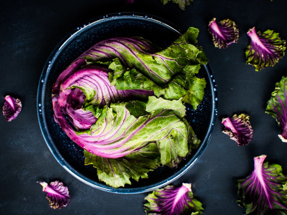 Top down view of a black stoneware bowl containing leafy greens with purple stems