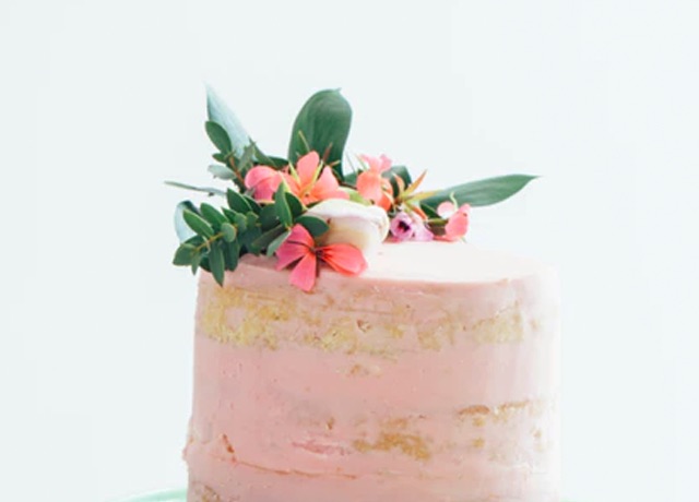 Pink iced cake topped with delicate pink florals and greenery, against a white background