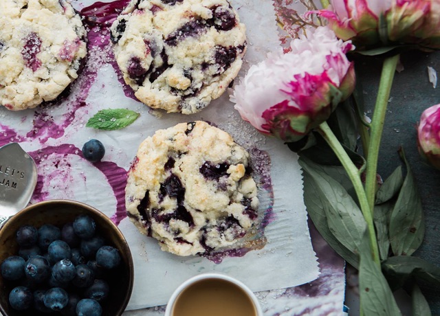 Top down view of berry scones on parchment paper, beside a bowl of blueberries and a couple cut pink and white flowers